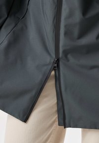 Giacca nera con cerniera a doppio senso parzialmente aperta sopra pantaloni beige, mostrando i dettagli della cerniera e la trama del tessuto.