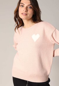 Pull léger rose en matière douce, avec un motif de cœur blanc sur la poitrine gauche et des bords côtelés aux poignets et à l'ourlet.