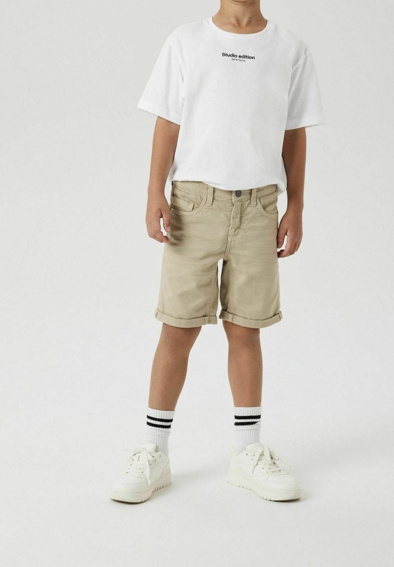 Enfant portant un T-shirt blanc avec texte, un short beige retroussé, des chaussettes blanches à rayures noires, et des baskets blanches sur un fond uni.