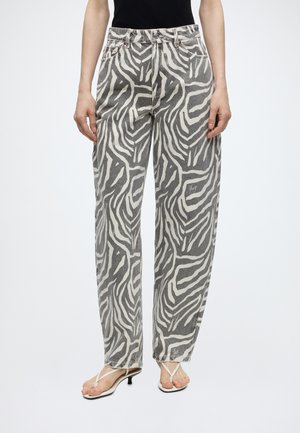 Person iført højtaljede, løstsiddende jeans med sort-hvid zebra mønster og hvide sandaler med hæl, stående mod en ensfarvet baggrund.