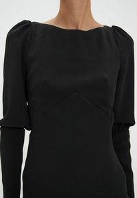 Robe noire avec décolleté carré, corsage structuré et manches bouffantes. Fabriquée en tissu lisse avec une silhouette ajustée.