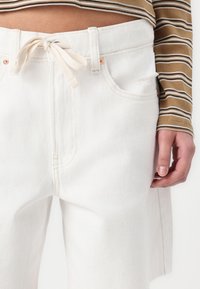 Pantalon en denim blanc avec taille haute et cordon de serrage, présentant des détails de coutures subtils, des poches latérales et une coupe décontractée.