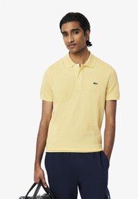 Polo in cotone giallo chiaro con colletto, tre bottoni e un logo ricamato verde sul lato sinistro del petto.