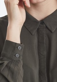 Chemise boutonnée verte olive avec un col classique, présentant une texture lisse et des poignets à deux boutons. Des plis subtils ajoutent du détail.
