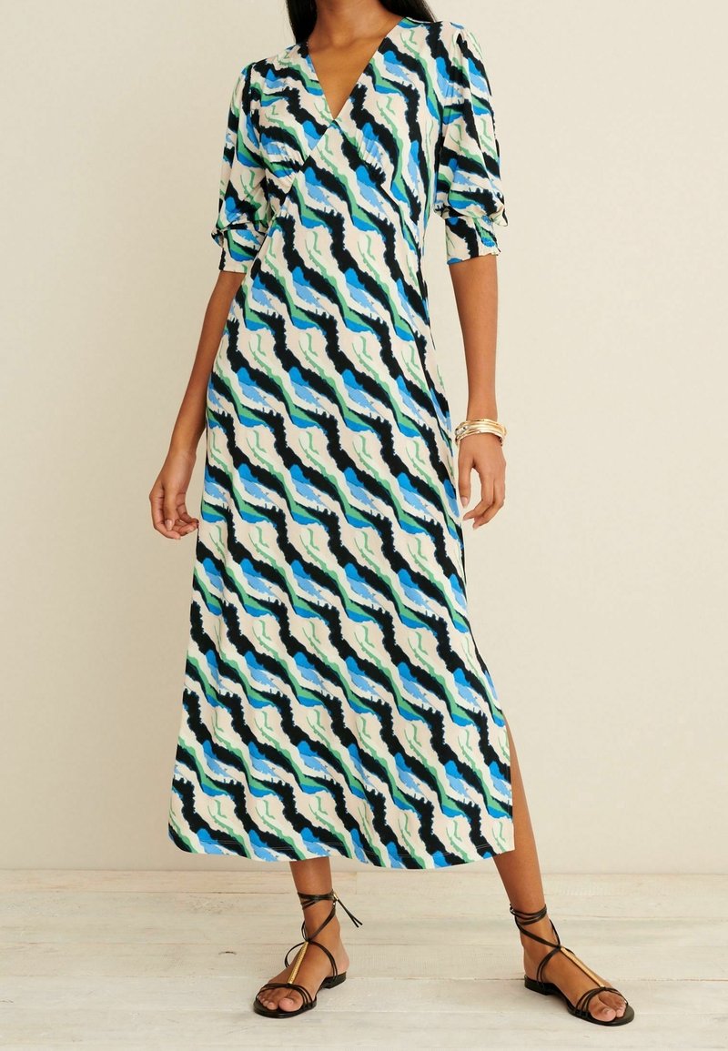 Femme portant une robe midi à imprimé abstrait coloré avec des manches trois-quarts bouffantes et des sandales noires à lacets, debout contre un fond uni.