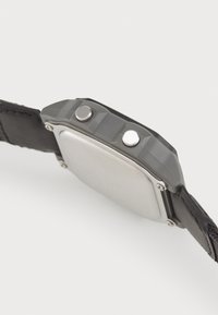 Montre rectangulaire avec un cadran en métal argenté et des accents gris, dotée de deux boutons et d'un bracelet en cuir noir avec un détail texturé.