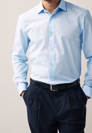 Mann trägt hellblaues langärmliges Hemd mit Knöpfen, in schwarze plissierte Hose mit schwarzem Gürtel gesteckt, Hände in den Taschen, vor neutralem Hintergrund.