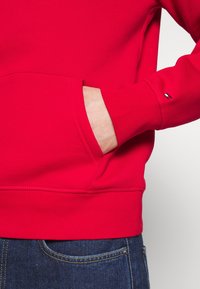 Mano in tasca anteriore di una felpa rossa con un piccolo logo sul polsino della manica, abbinata a jeans in denim blu.