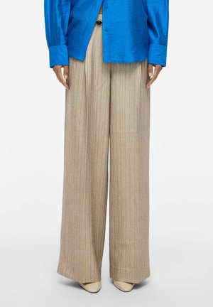Pantalon beige à jambes larges avec fines rayures, associé à une chemise bleu vif et des chaussures pointues beiges sur fond blanc.
