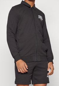 Chaqueta negra con cremallera hecha de una mezcla de algodón, con cuello, mangas largas y un logo en el pecho, combinada con pantalones cortos a juego.