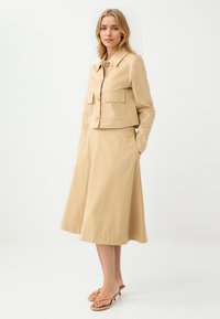 Veste beige courte avec deux poches plaquées et fermeture à bouton, associée à une jupe midi beige. Le mannequin porte des sandales à lanières beiges.