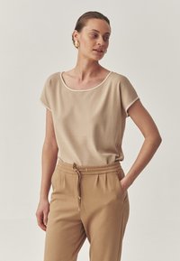 Bluză bej cu mânecă scurtă, decolteu rotund și accente albe, purtată cu pantaloni din material moale cu șnur maro. Material neted, croi lejer.