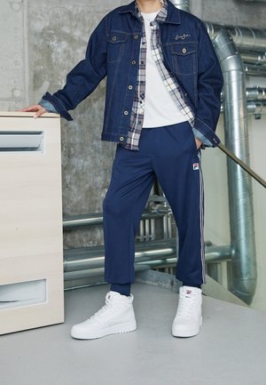 Denimjakke med guldhardware, lagd over en ternet skjorte, parret med marineblå sportsbukser med sidelister og hvide sneakers.