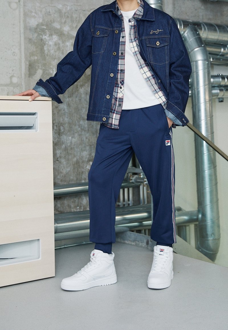 Veste en denim avec des ornements dorés, superposée à une chemise à carreaux, associée à un pantalon de sport bleu marine avec des bandes latérales, et des baskets blanches.