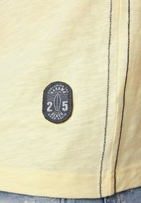 Camisa amarilla clara de mangas cortas con un acabado texturizado, que presenta un parche redondo de color azul oscuro con gráficos de surf y texto blanco a lo largo del dobladillo.