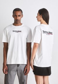 Camiseta de algodón blanca con cuello redondo, que presenta el logo "Tommy Jeans" en rojo y azul en la parte delantera y trasera. Diseñada con corte recto.