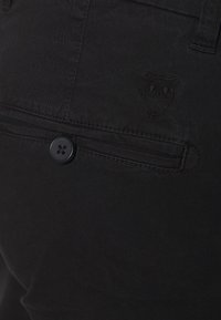 Svart byxor gjorda av bomullsblandning, med en bakficka med en svart knapp och subtil broderad ugglelogotyp. Slät textur med en figursydd passform.