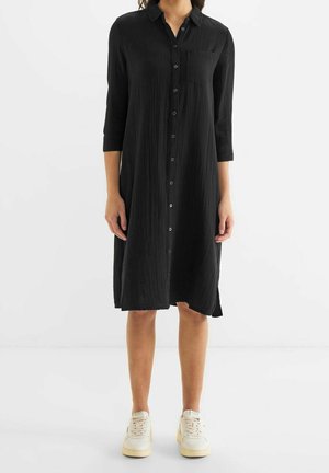 Black button-up dress with three-quarter sleeves, textured fabric, single chest pocket, and a loose fit, paired with white sneakers.
