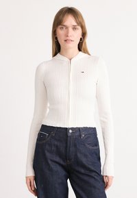 Cardigan bianco a costine con zip e colletto tondo. Abbinato a jeans in denim blu scuro a vita alta con tasche frontali.
