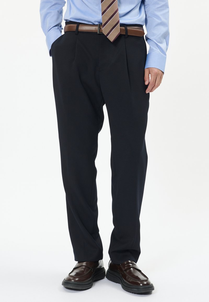 Tmavě modré plisované kalhoty se slim fit, doplněné hnědým koženým opaskem a lesklými hnědými botami. Viditelný detail texturované tkaniny.