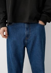 Modré denimové džínsy s klasickým strihom, s jednou prednou kapsou, viditeľným stehom a mierne štruktúrovaným povrchom. Skombinované s čiernym topom s dlhým rukávom.