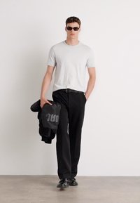 T-shirt gris à col rond, pantalon noir sur mesure, chaussures noires et lunettes de soleil ; tenant une veste bomber noire avec un détail de logo audacieux.