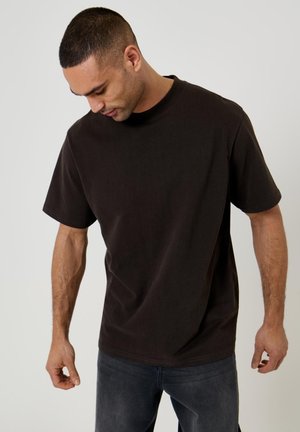 Uomo che indossa una T-shirt nera a maniche corte semplice e jeans scuri, che guarda verso il basso con le braccia rilassate lungo i fianchi.