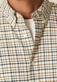 Geruite shirt met een beige, crème en bruine patroon, met groene en marineblauwe accenten. Knop-down kraag en zichtbare knoopdetails.