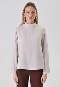 Langärmlige Bluse, weicher hellbeiger Stoff, hoher Ausschnitt, lockere Passform, glatte Textur, ergänzt durch dunkelbraune Hosen.