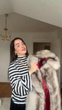 Nő fekete-fehér csíkos felsőben, aki egy szürke szőrmebundát tart piros béléssel, egy csillárral rendelkező szobában.