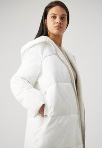 Doudoune blanche avec une capuche duveteuse et une doublure douillette, présentant une fermeture à glissière à l'avant, des poches latérales et une texture matelassée.