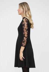 Vestido negro con mangas largas de encaje transparente con motivos florales, corpiño ajustado, falda en línea A y textura suave. Diseño minimalista.