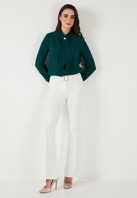 Teal blouse with a bow tie collar and button closure, paired with white flared trousers and silver snakeskin heels. Smooth fabric, clean design.