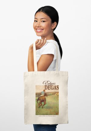 Usměvavá žena drží béžovou tašku s potiskem obrazu Edgara Degase "Džokej v modrém na kaštanovém koni".