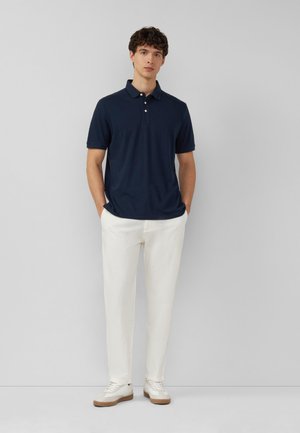 Mężczyzna stoi przy szarej ścianie w granatowej koszulce polo z krótkim rękawem, białych spodniach i białych trampkach z gumową podeszwą, ręce w kieszeniach.