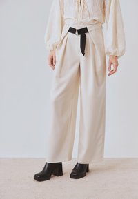 Los pantalones anchos de color crema presentan pliegues en la parte frontal y un cinturón negro con una hebilla circular. Se combinan con botas negras de tacón.