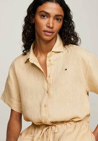 Abito camicia in lino beige con abbottonatura frontale, maniche corte e logo ricamato sul petto. Presenta una vita con cordoncino.