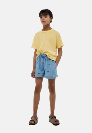Enfant debout, les mains dans les poches, portant un t-shirt jaune, un short bleu clair brodé et des sandales marron sur fond blanc.