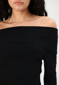 Svart off-shoulder ribbestrikket topp med bred halsutringning og lange ermer, laget av et teksturert, elastisk stoff.