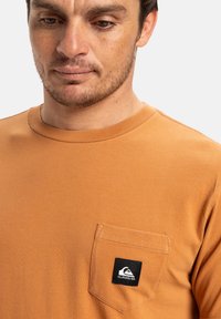 Oranžové bavlněné tričko s kulatým výstřihem, vybavené náprsní kapsou a černým logem. Látka vypadá hladce a má jednobarevný design.