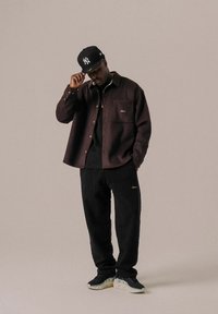 Uomo che indossa una camicia scura con bottoni, pantaloni sportivi neri, sneaker e un cappellino da baseball nero, in piedi davanti a uno sfondo beige semplice, toccando il bordo del cappello.