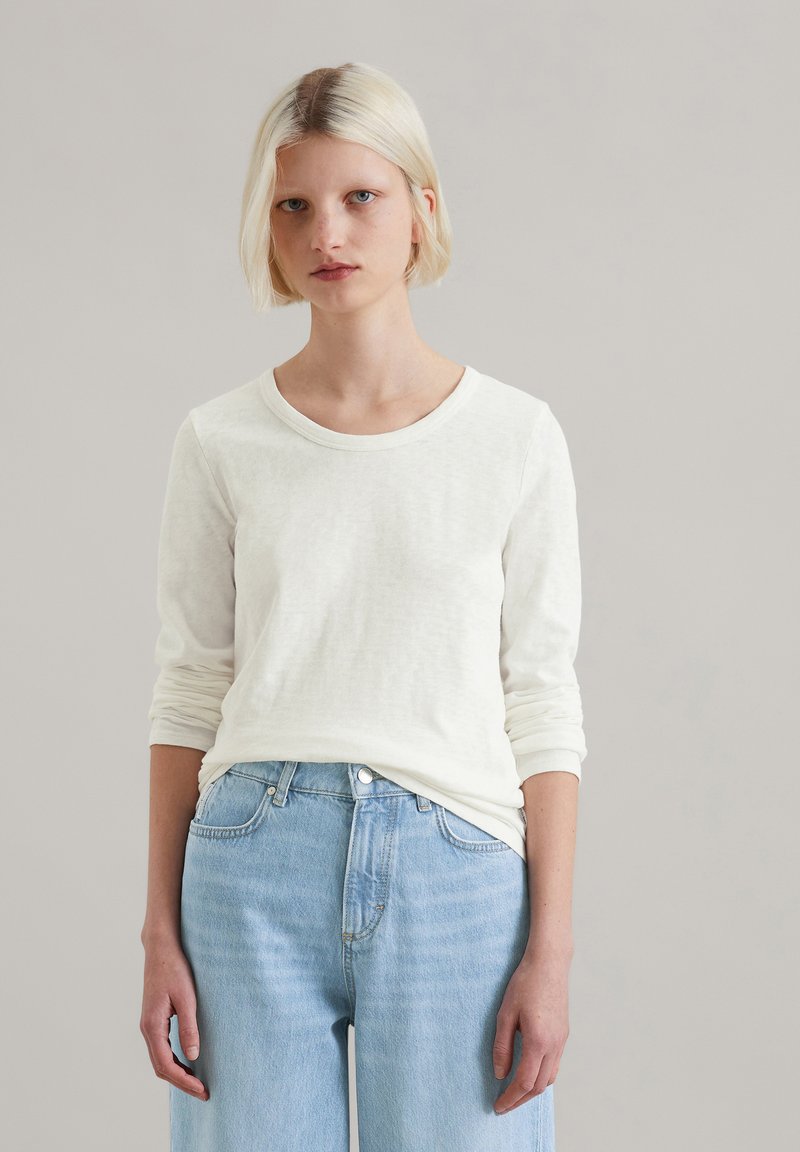 Koszula z długim rękawem i okrągłym dekoltem w kremowym kolorze, wykonana z teksturowanej tkaniny, zestawiona z jasnoniebieskimi, luźnymi dżinsami.