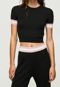 Žena v černém žebrovaném topu a kalhotách se sadou růžových a bílých pruhů na rukávech a pasu, odhalující midriff.