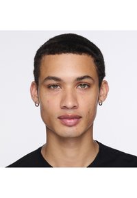 Potret seseorang dengan rambut keriting gelap pendek dan mata hijau muda, mengenakan anting hoop kecil dan kaos hitam, dengan kulit yang bersih.