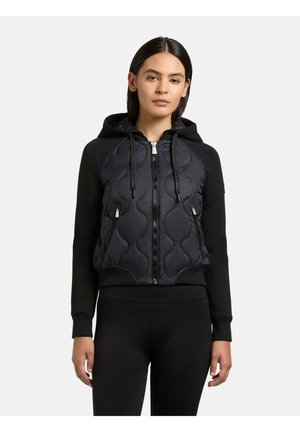 Kvinde iført en sort quiltet hættetrøje med lynlås, snore og lynlåslommer, kombineret med sorte tætsiddende bukser, stående foran en hvid baggrund.