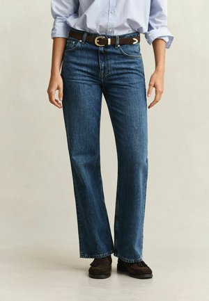 Personne portant un jean bleu droit, une ceinture marron à boucle dorée, une chemise bleu clair retroussée et des mocassins marron foncé.