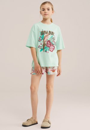 Jeune fille debout en t-shirt oversize vert clair avec imprimé oiseaux et fleurs, short à motifs et chaussures beiges sans lacets.