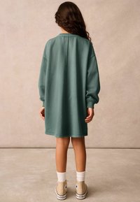 Prevelika zelena obleka iz sweatshirt materiala z dolgimi rokavi, rebrastimi manšetami in okroglim izrezom, v kombinaciji s bež supergami in belimi nogavicami.