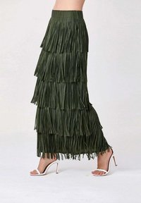Jupe maxi à franges vert olive avec des couches superposées, présentant une finition texturée. Associée à des sandales à talons hauts blanches.