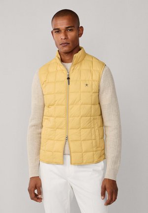 Homme portant un gilet matelassé jaune sans manches par-dessus un pull en tricot beige et un pantalon blanc, debout devant un fond gris clair uni.